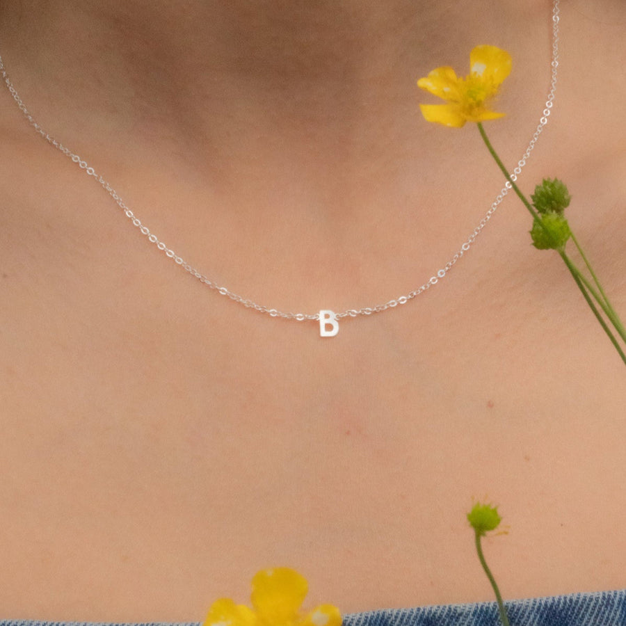 Person wearing a silver necklace with a 'B' pendant, surrounded by yellow flowers.