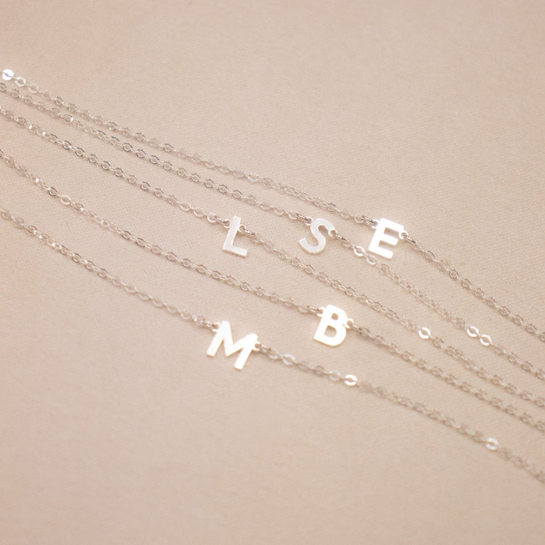 Three silver necklaces with letter pendants on a beige background