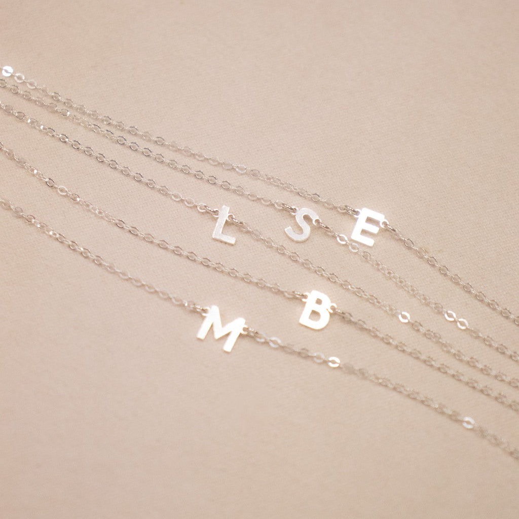 Three silver necklaces with letter pendants on a beige background