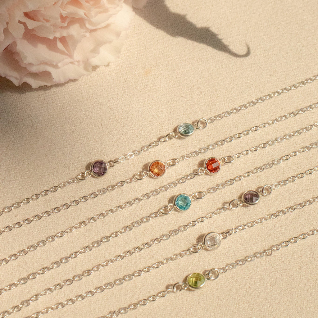 Set of silver bracelets with colored gemstones on a sandy surface with a pink flower and green leaves.