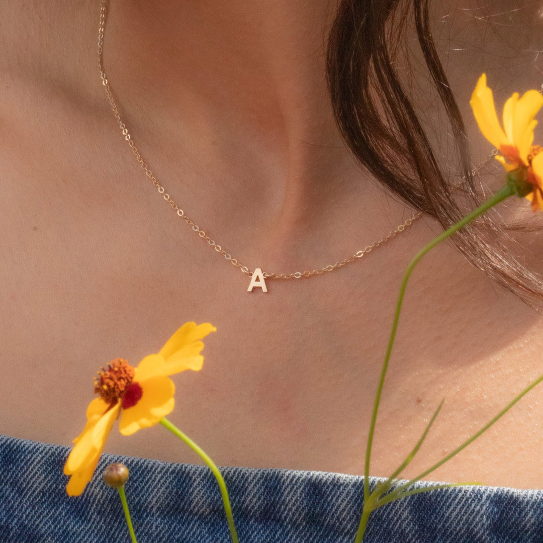 Necklace with a letter 'A' pendant worn by a person, with yellow flowers in the foreground.