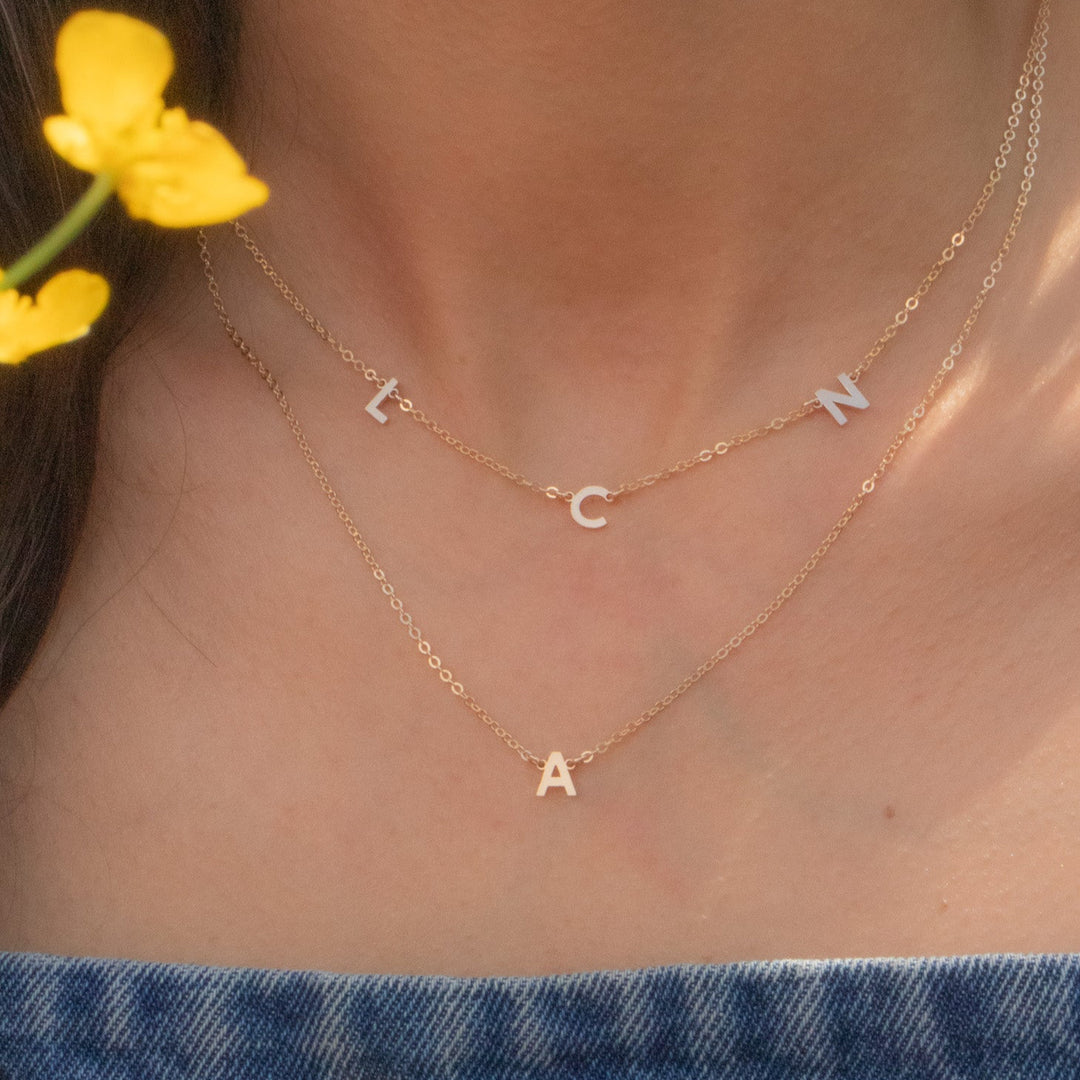 Close-up of a person wearing gold necklaces with letter pendants, surrounded by yellow flowers.