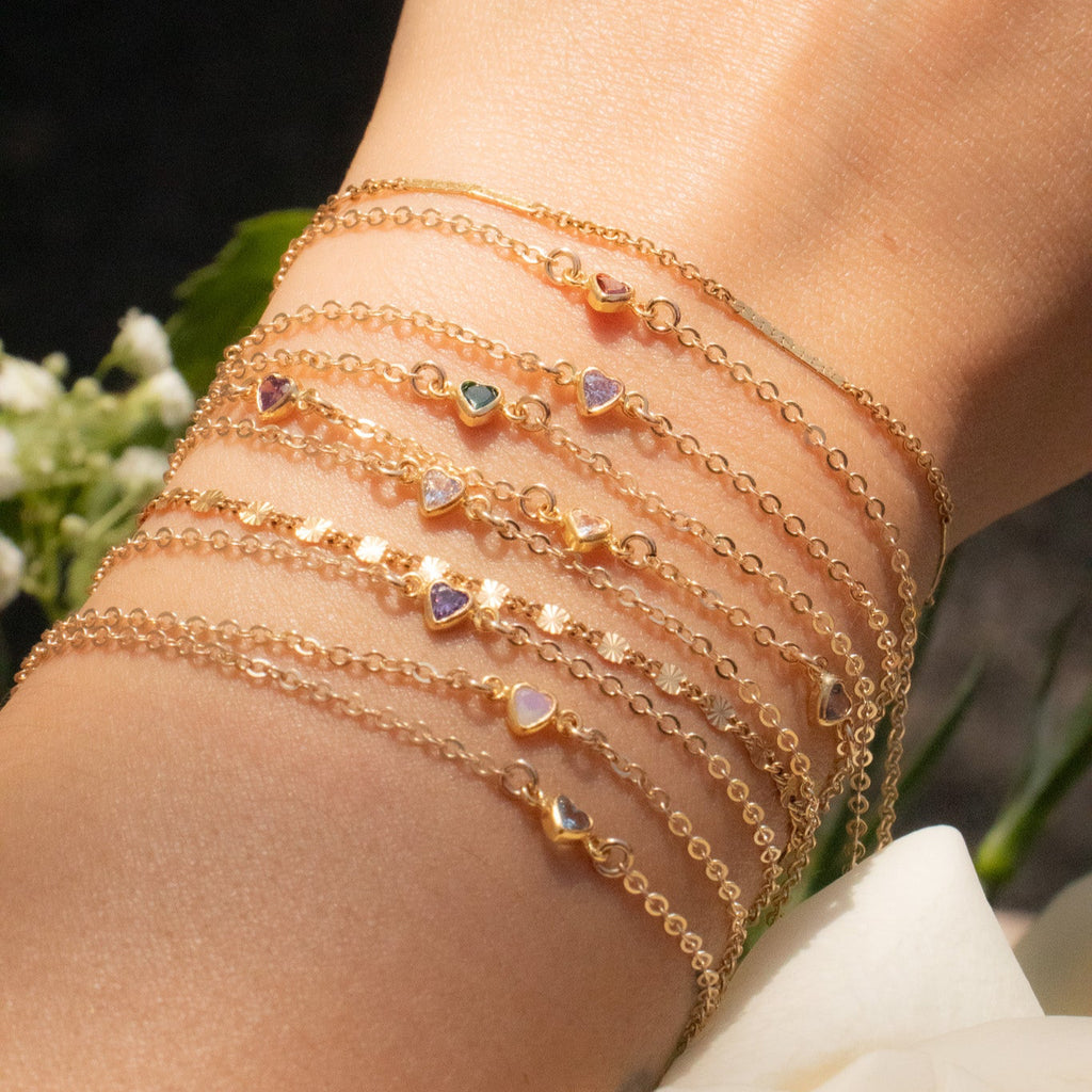 Close-up of a wrist wearing multiple gold bracelets with heart charms, against a blurred floral background.