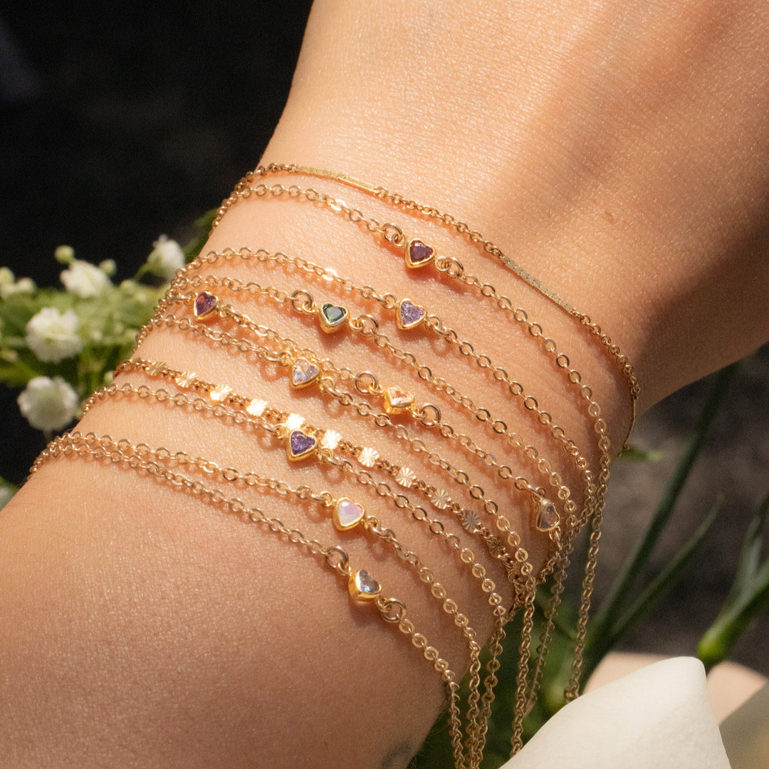 Close-up of a wrist wearing multiple gold bracelets with small heart charms, against a blurred natural background.