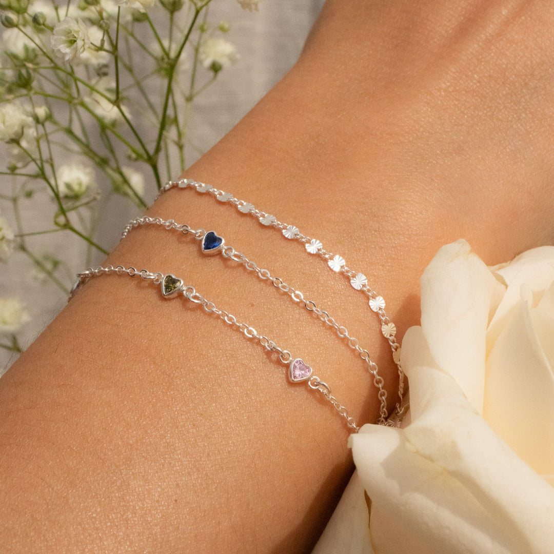 Close-up of a wrist wearing multiple silver bracelets with heart charms, against a blurred floral background.