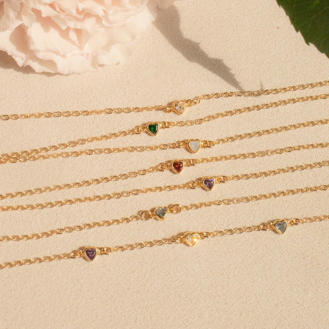 Gold bracelets with heart-shaped gemstones on a beige surface with a flower and leaves in the background.