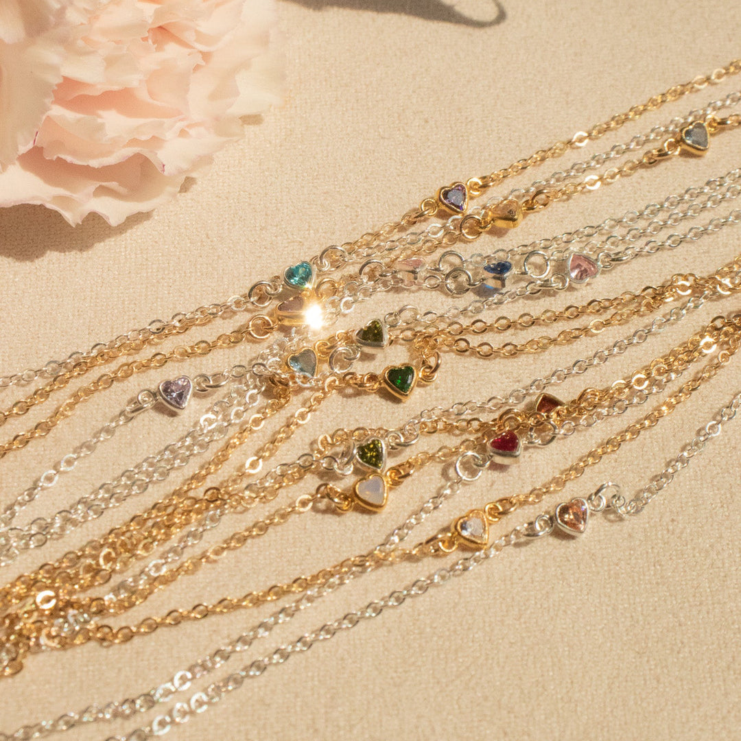 Multiple gold heart birthstone bracelets with gemstones on a beige surface