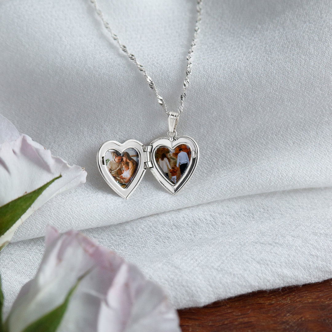 Silver heart-shaped locket with two photo panels on a white fabric background with a white flower.