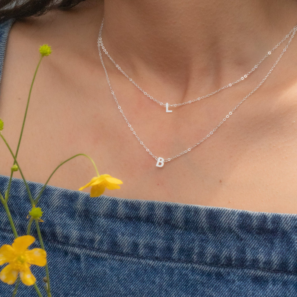 Necklace with letter 'B' pendant worn by a person, with denim fabric and yellow flowers in the foreground.