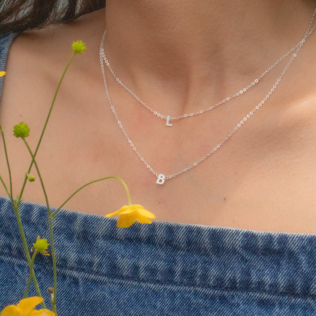 Close-up of a person wearing two silver necklaces with letters, surrounded by yellow flowers and denim fabric.