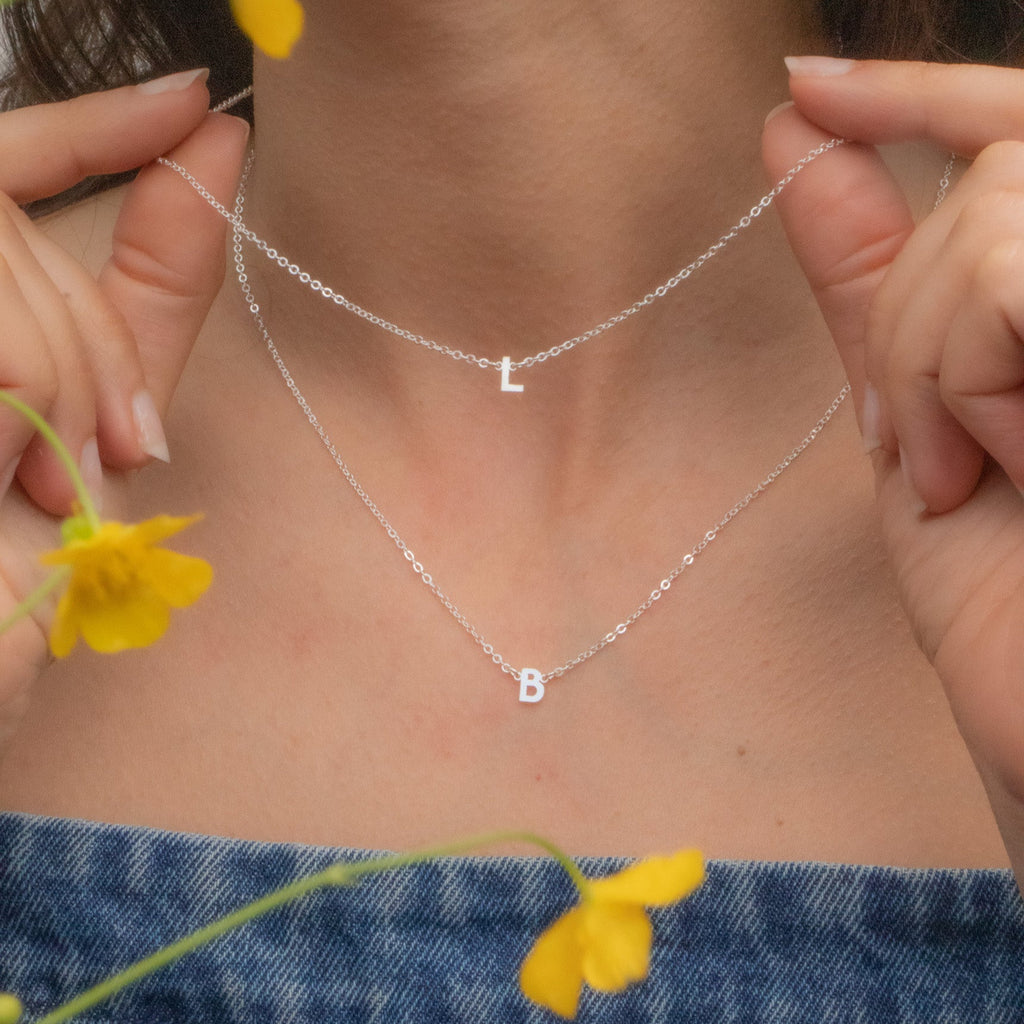 Person wearing a silver necklace with letter 'B' pendant, holding yellow flowers, wearing blue denim.