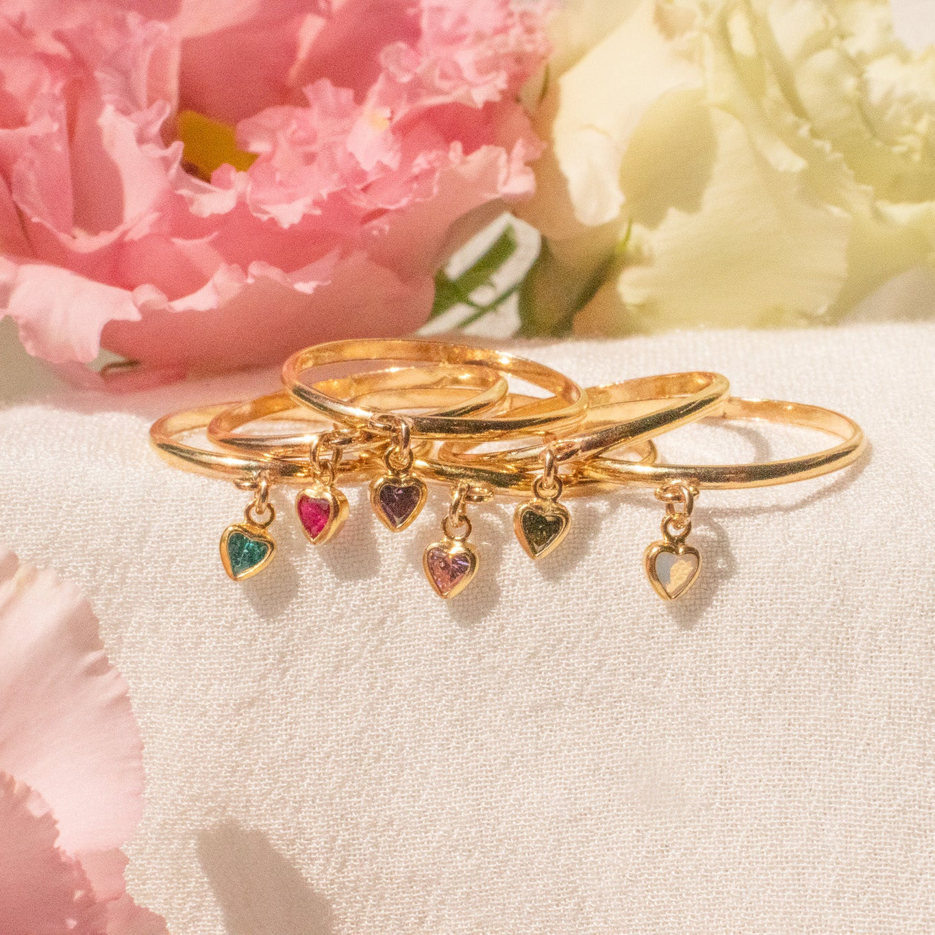 Gold rings with heart-shaped charms on a white surface with pink flowers in the background