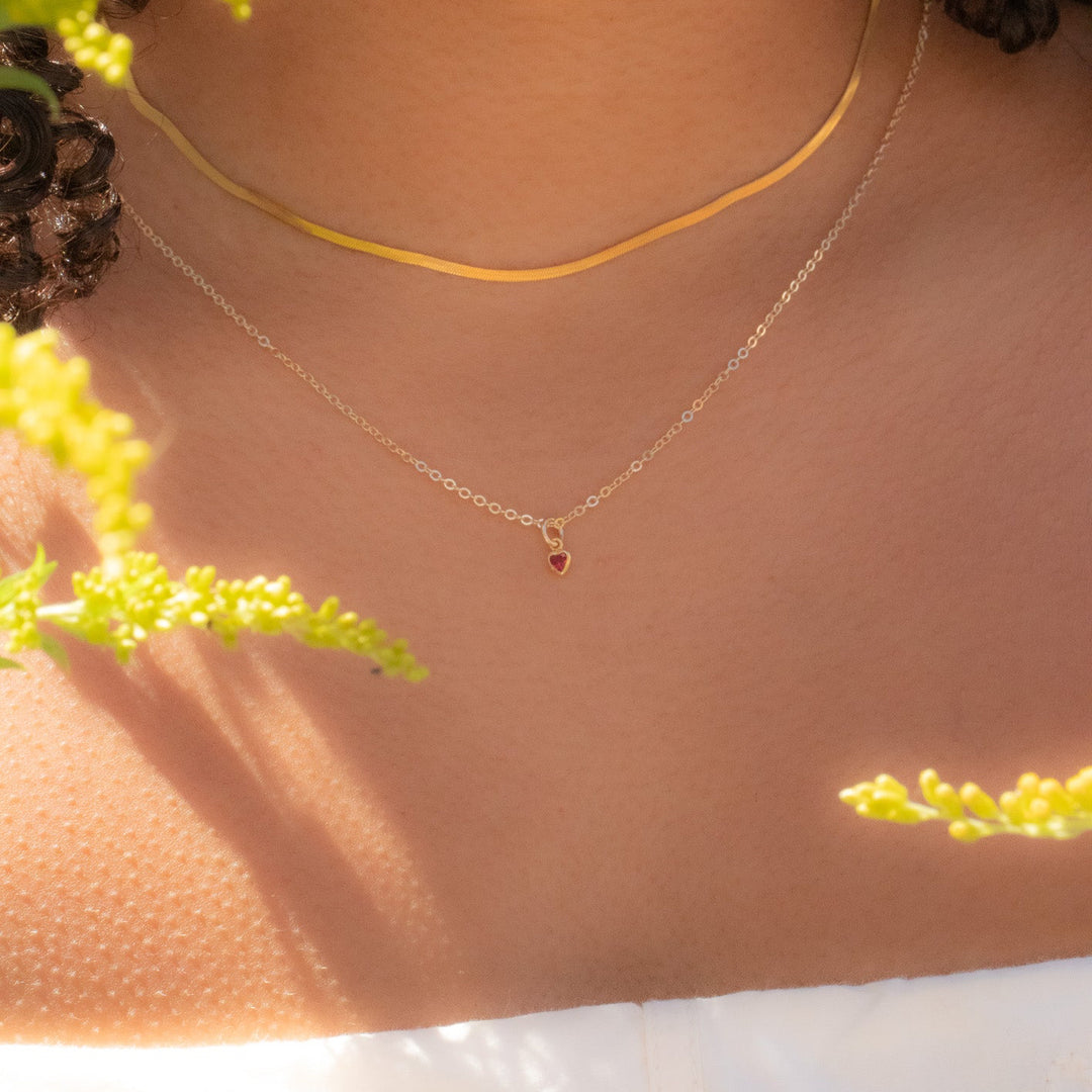 Close-up of a person wearing a gold necklace with a small red pendant, surrounded by yellow flowers.